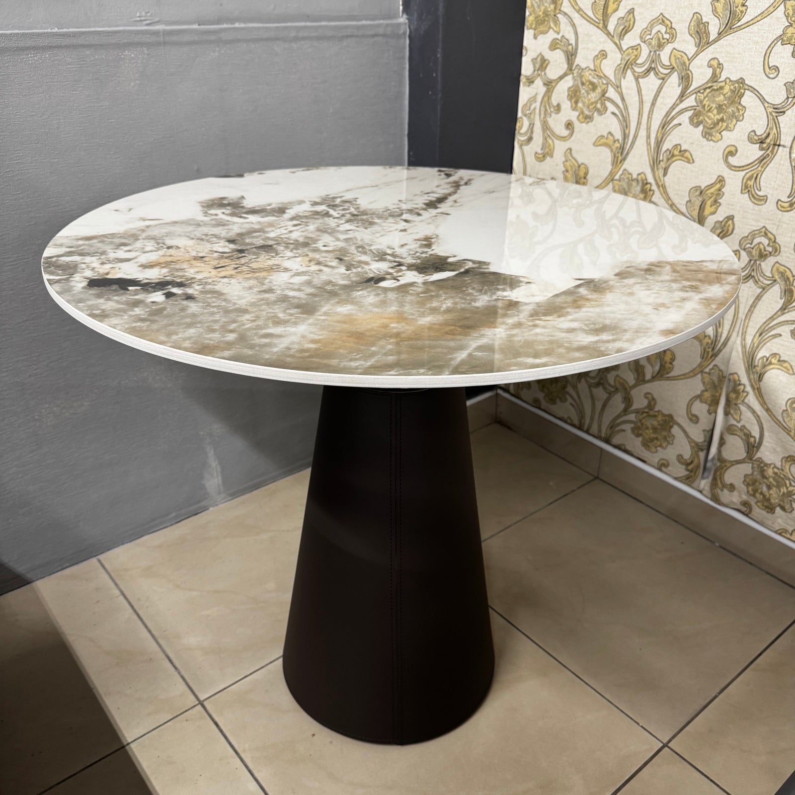 A round marble-top table with a dark tapered pedestal base, shown in an indoor showroom with patterned wallpaper and tiled floor.