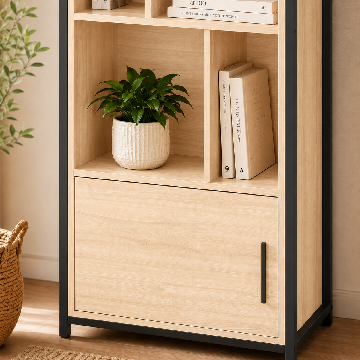 A light natural wood bookcase with black metal frame and legs, featuring open shelves on top and a single drawer with a black handle below. The bookcase is styled with books and a potted green plant, set in a warm indoor room environment.