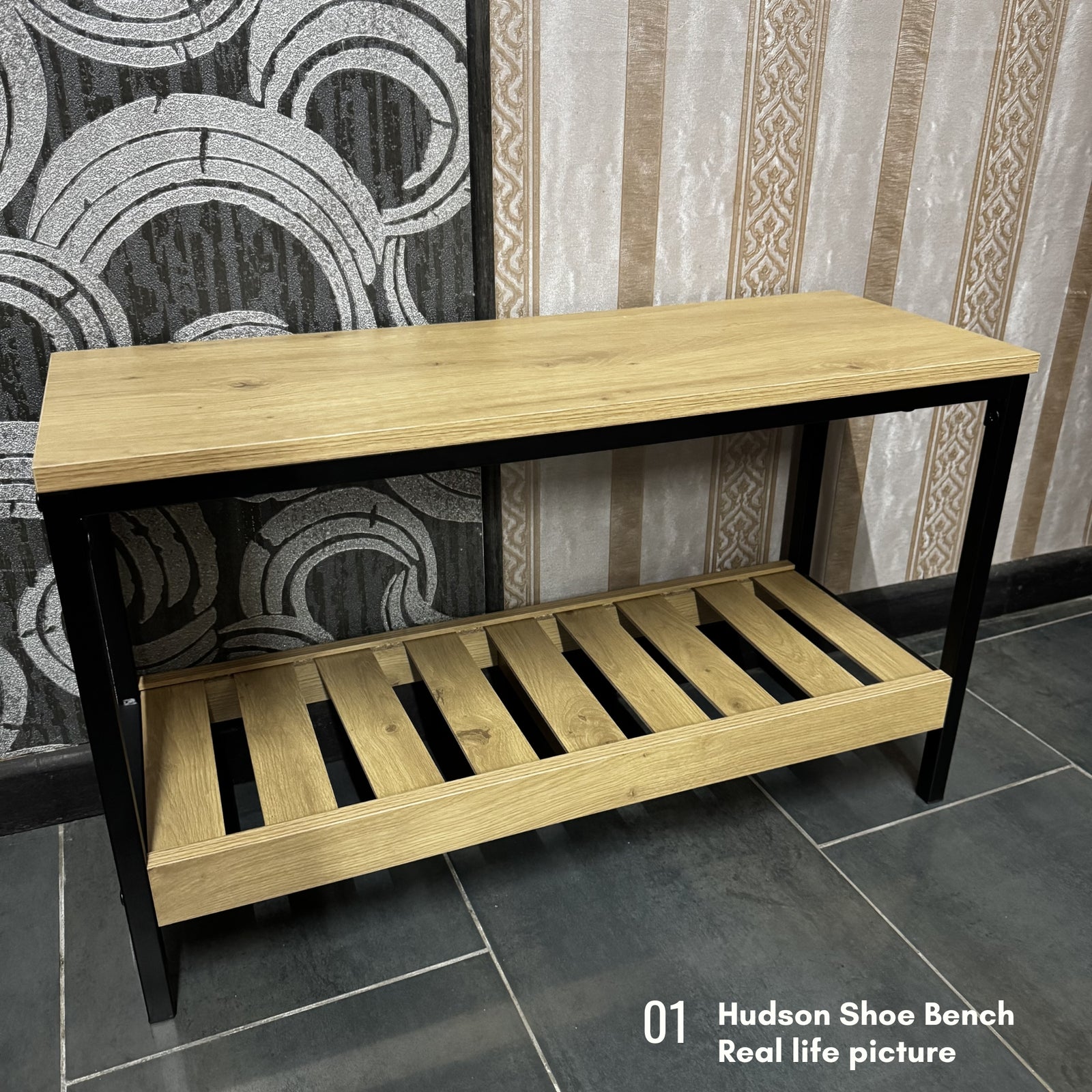 A wooden shoe bench with a light oak top and slatted lower shelf, supported by a black metal frame, shown in a real interior setting with patterned wallpaper and tiled floor. Text in the corner reads '01 Hudson Shoe Bench Real life picture'.