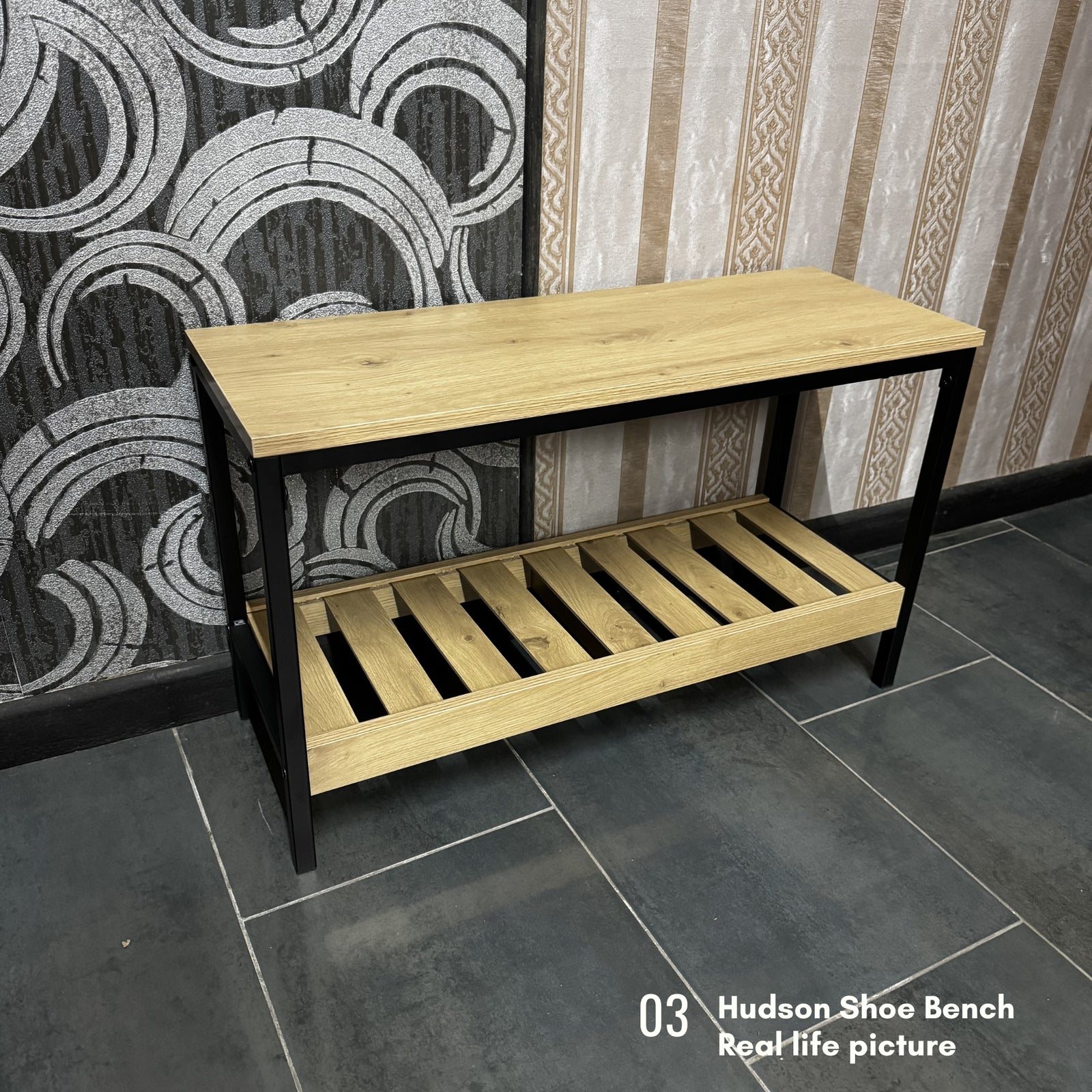 A wooden shoe bench with a light oak top and slatted lower shelf on a black metal frame, shown in a real interior setting with patterned wallpaper and dark tile flooring. Text in the lower right reads '03 Hudson-Shoe Bench Real life picture'.