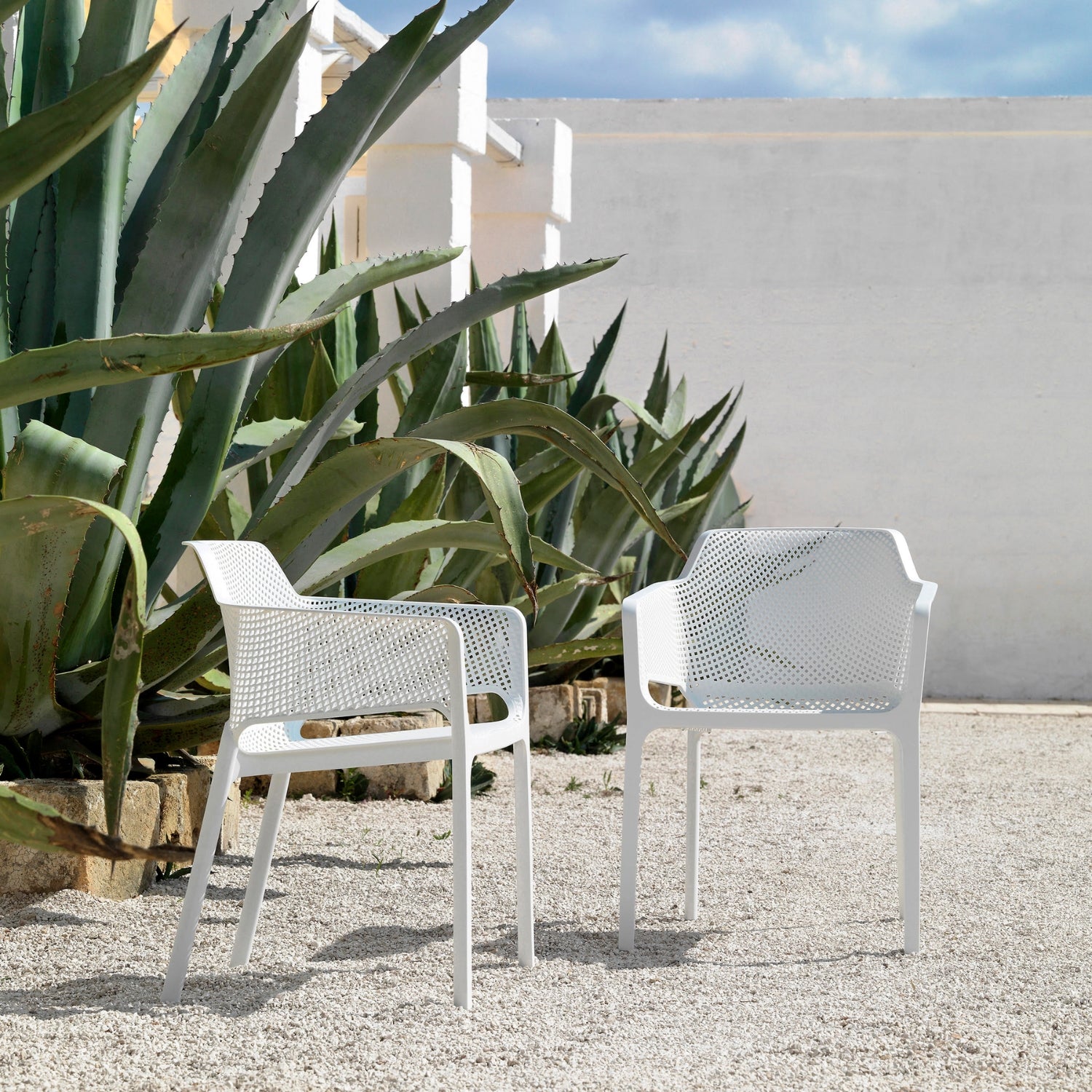 Two white Replica Net Armchairs placed in a modern garden with large plants, ideal for outdoor relaxation.