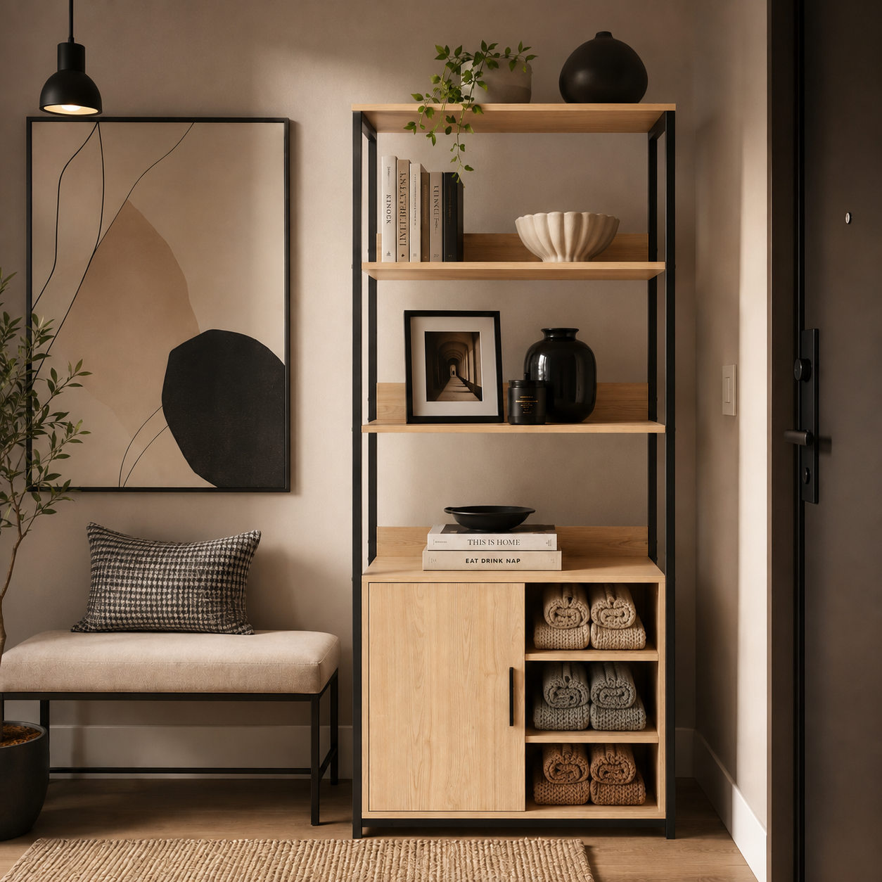 A tall open shelving unit with three light wood shelves and a lower cabinet section, styled with books, vases, a bowl, and folded towels. It is placed in a warm, modern entryway or home office setting next to a bench and a framed abstract print.