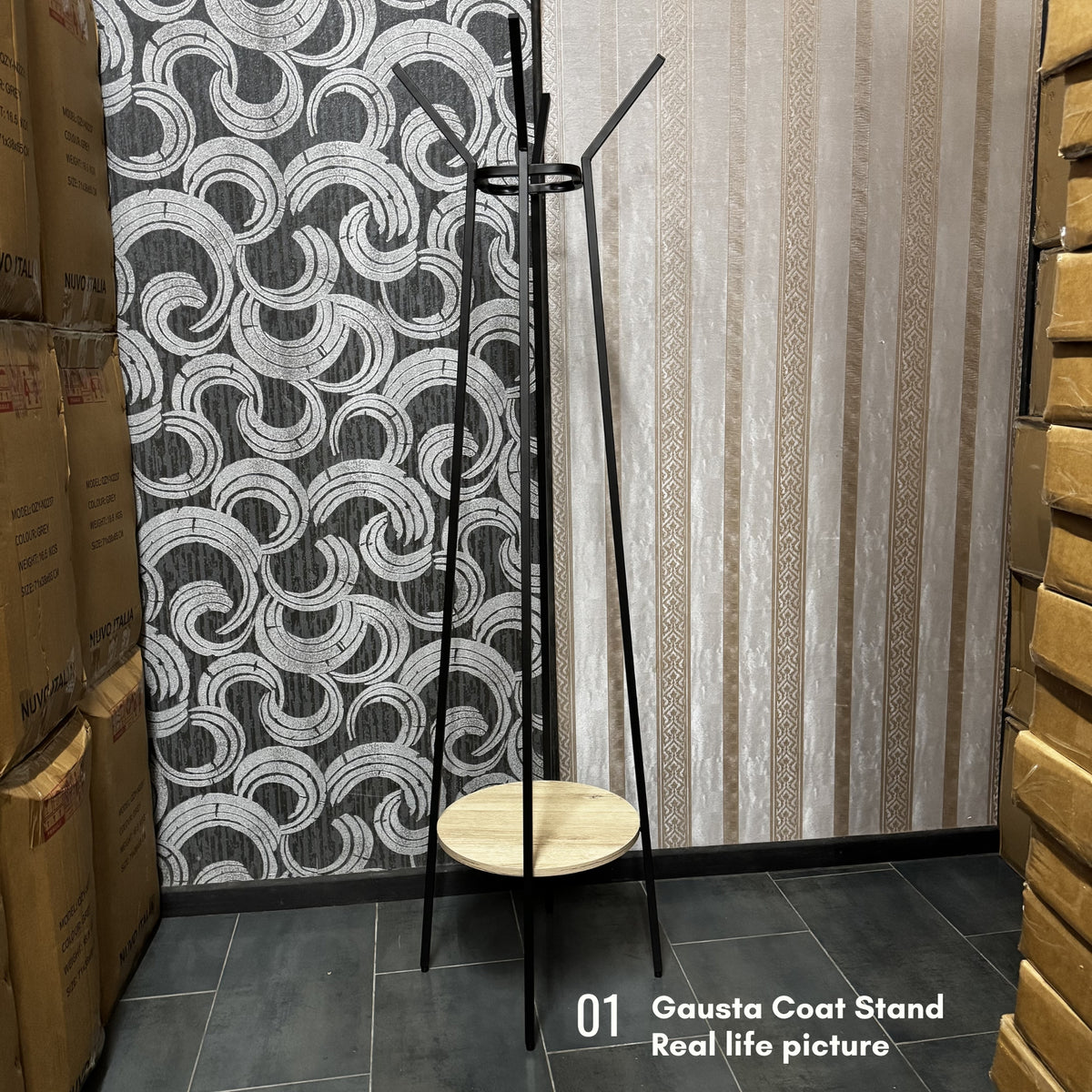 A black metal coat stand with a round light wood seat and backrest, shown in a warehouse-like room with patterned wallpaper, stacked cardboard boxes, and a text overlay reading '01 Gausta Coat Stand Real life picture'.