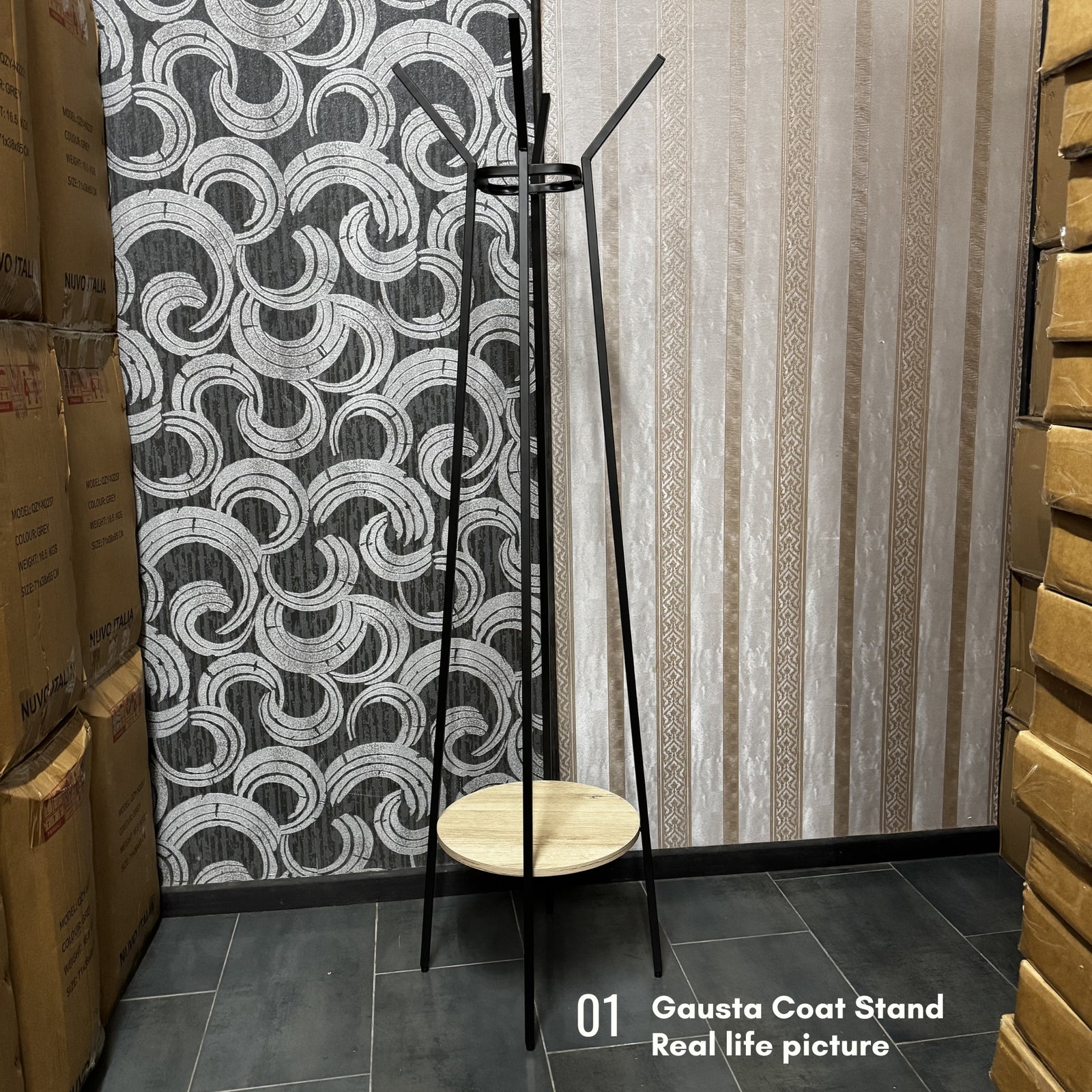 A black metal coat stand with a round light wood seat and backrest, shown in a warehouse-like room with patterned wallpaper, stacked cardboard boxes, and a text overlay reading '01 Gausta Coat Stand Real life picture'.