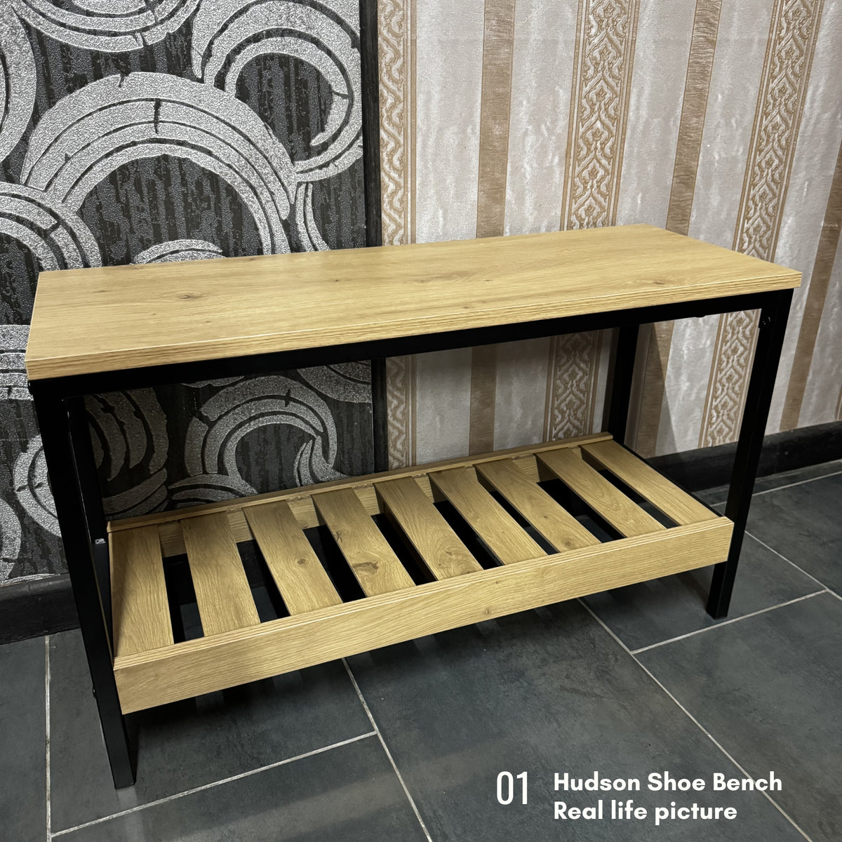 A wooden shoe bench with a light oak top and slatted lower shelf, supported by a black metal frame, shown in a real interior setting with patterned wallpaper and tiled floor. Text in the corner reads '01 Hudson Shoe Bench Real life picture'.