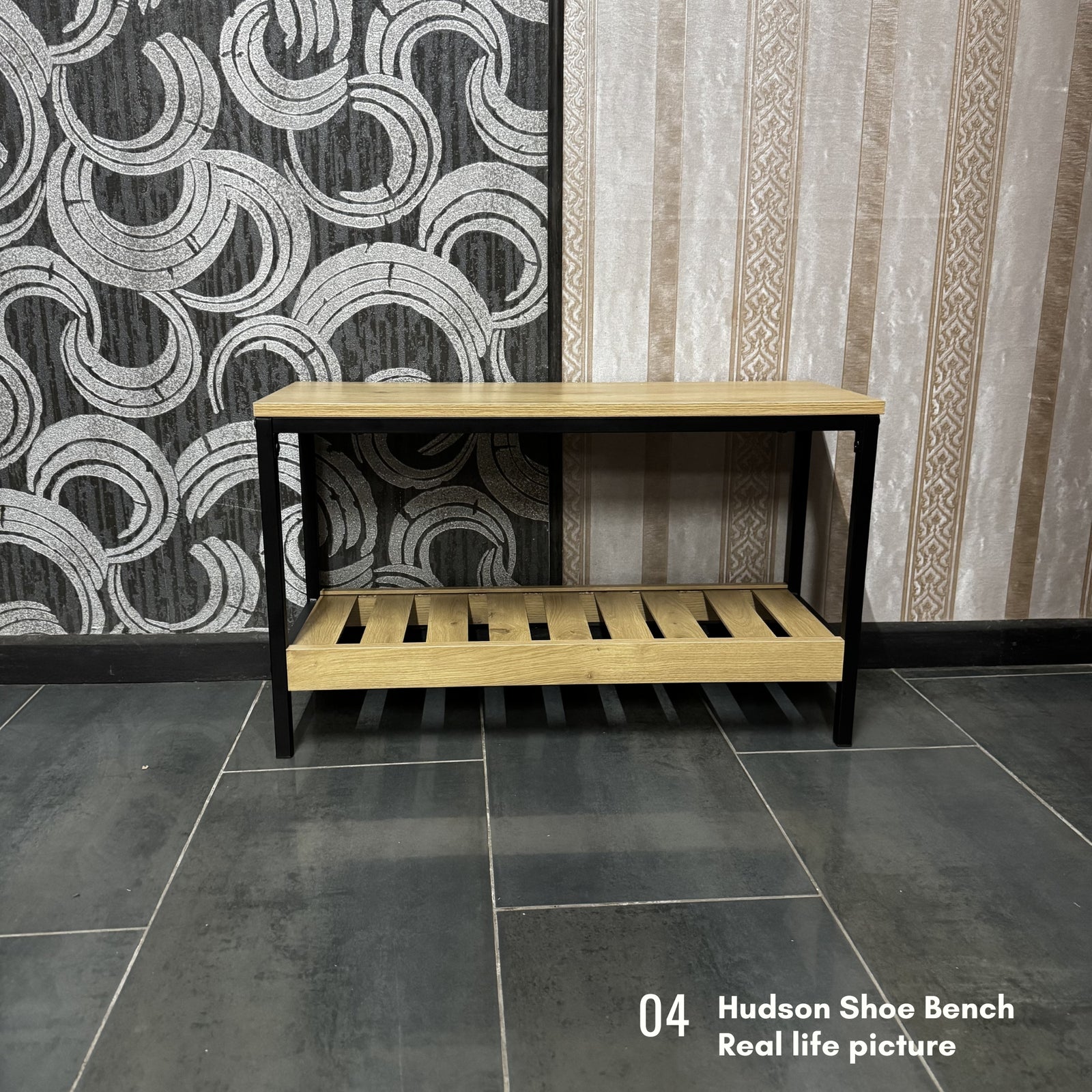 A wooden shoe bench console table with a light natural wood top and slatted lower shelf, supported by a black metal frame, set against patterned wallpaper and dark tiled flooring.