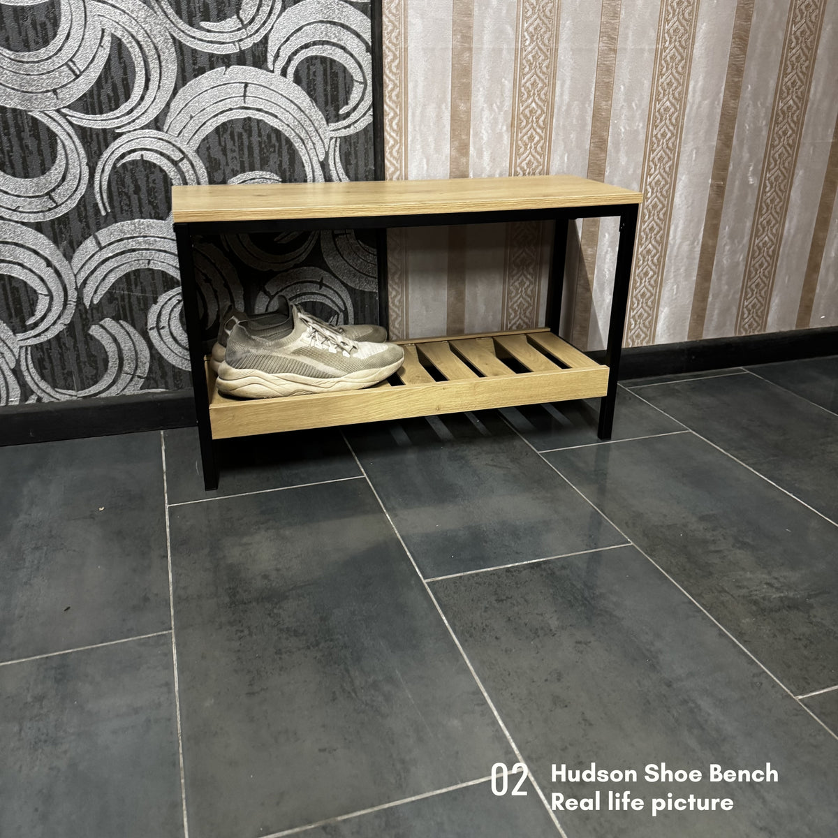 A two-tier wooden shoe bench with a light oak top and shelf, black metal frame, and a pair of white sneakers on the lower shelf, set against a patterned wall and dark gray tiled floor.