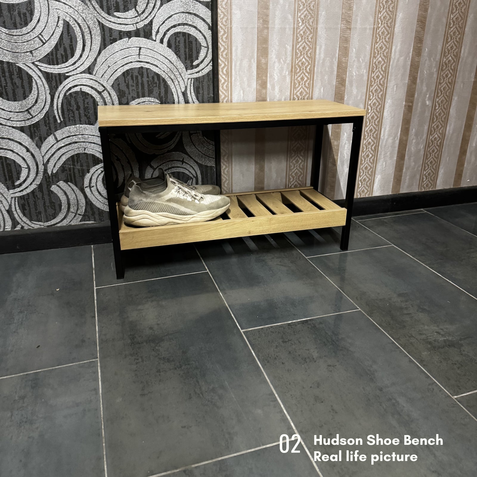 A two-tier wooden shoe bench with a light oak top and shelf, black metal frame, and a pair of white sneakers on the lower shelf, set against a patterned wall and dark gray tiled floor.