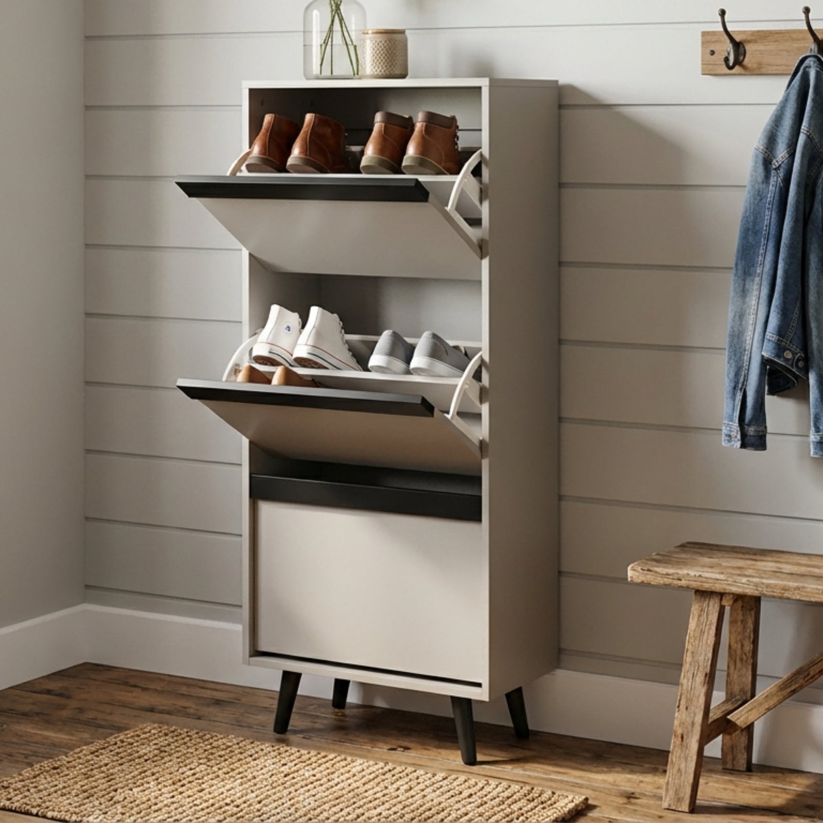 A tall, narrow shoe cabinet in a light gray finish with black legs and open shelves, shown holding several pairs of shoes in a styled room with a wood floor, shiplap wall, stool, and hanging jacket.