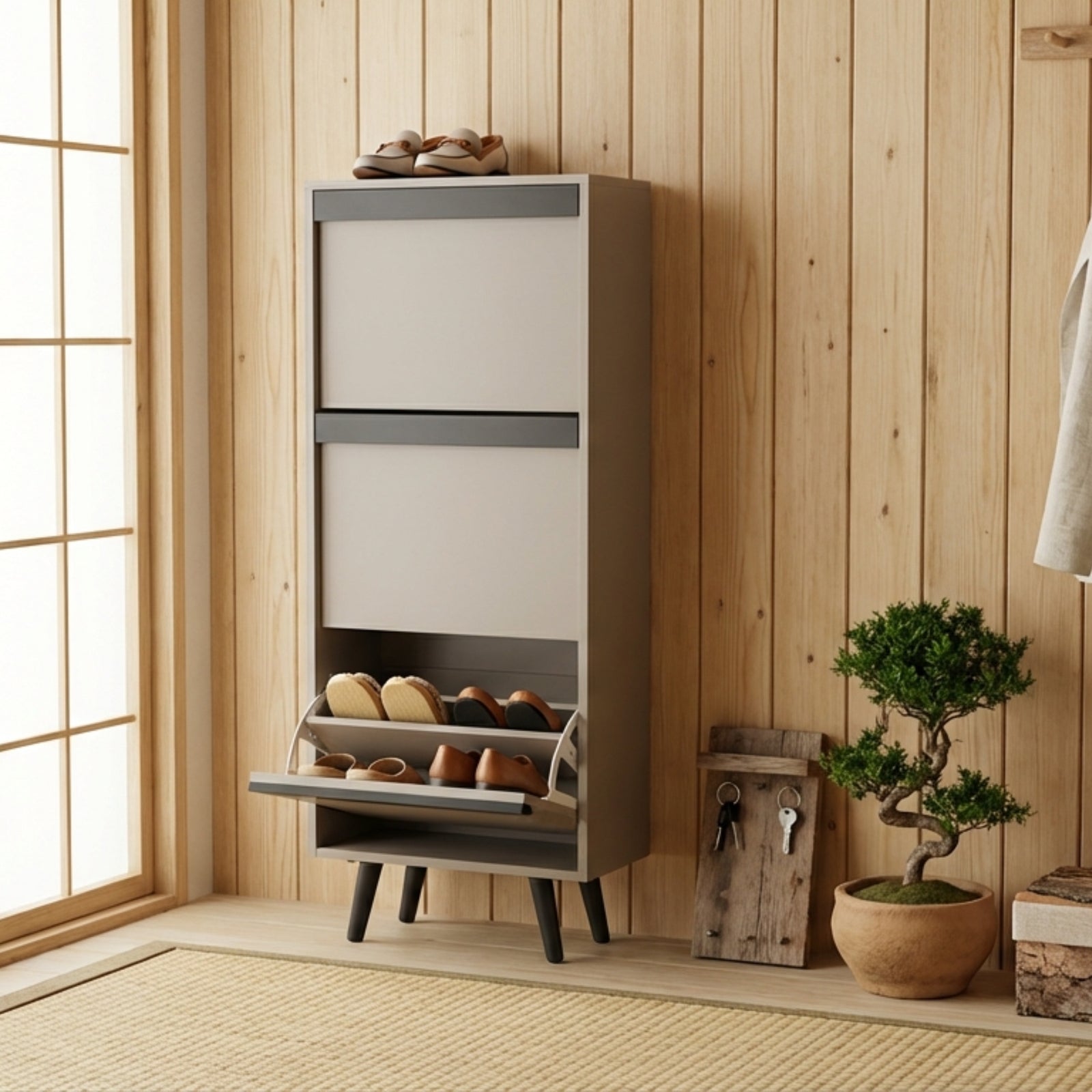 A tall, narrow shoe cabinet in a warm gray and beige finish stands against a light wood-paneled wall in a Japanese-style room. The lower drawer is open, showing several shoes inside, and a pair of sandals rests on top.